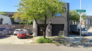 A tree stands next to a building in an outdoor setting, with a parking lot with various vehicles .