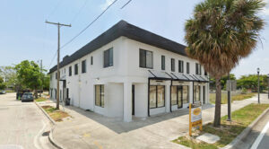 A building with a black roof surrounded by trees and a clear sky. The structure features windows and is located in a residential area, with a porch visible. There are palm trees nearby and a road in front of the property