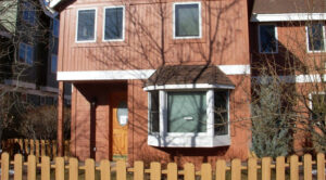 Exterior view of a brown wooden house with a bay window, surrounded by a picket fence and bare trees, on a sunny day.