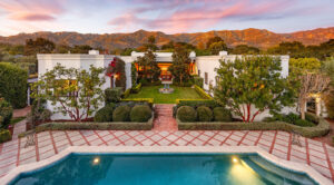 Stunning modern home surrounded by lush gardens and mountains, featuring a pool in the foreground and vibrant sunset sky.