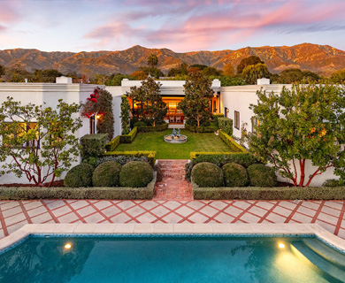 Stunning modern home surrounded by lush gardens and mountains, featuring a pool in the foreground and vibrant sunset sky.